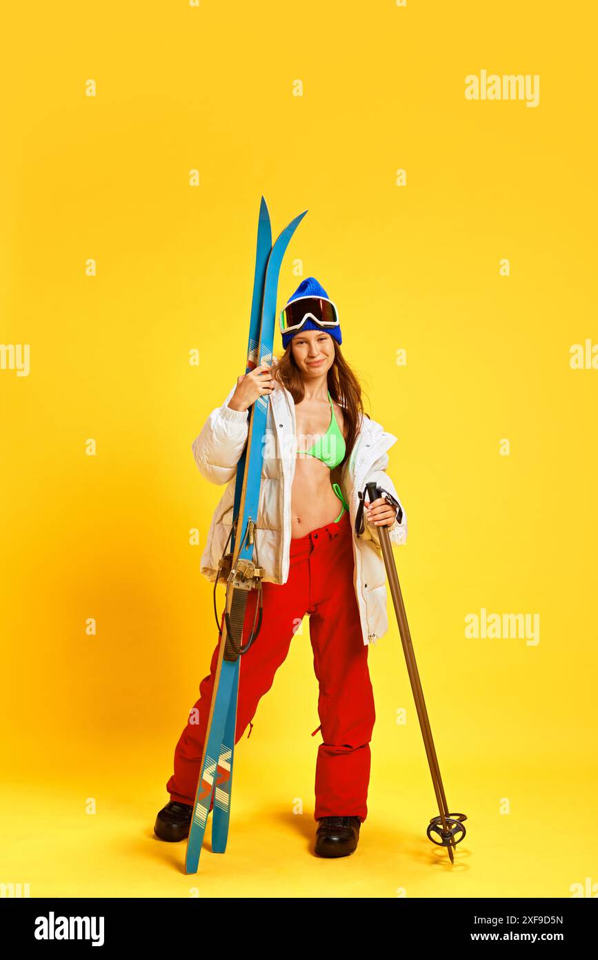 Ragazza che combina abbigliamento estivo e invernale, tenendo in mano l'attrezzatura da sci, posato su uno sfondo giallo brillante Foto Stock