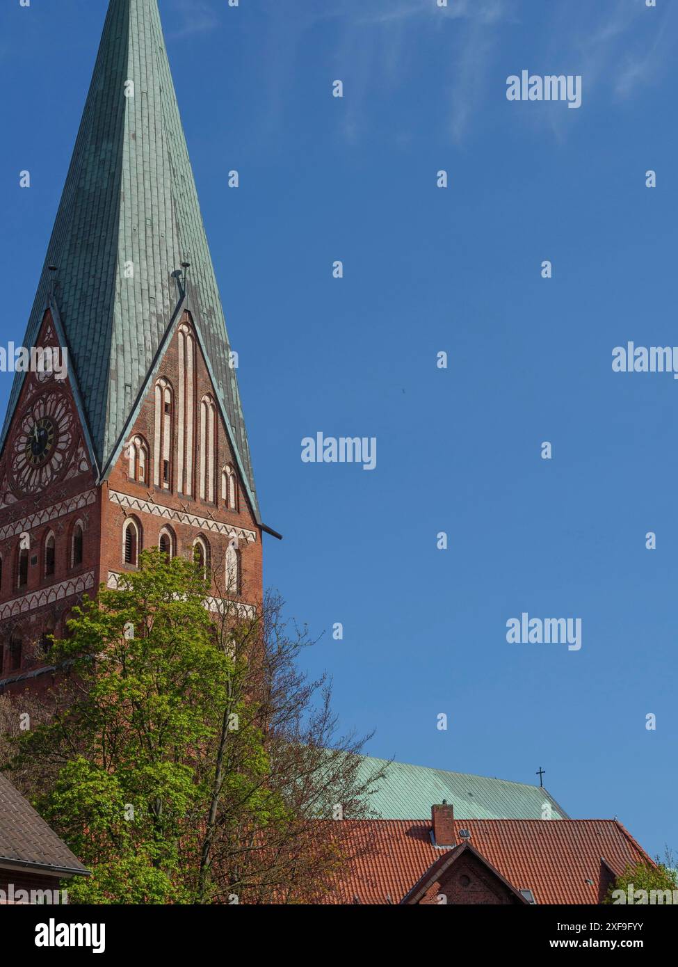 Una torre di chiesa con una guglia e un edificio rosso mattoni sono parzialmente oscurati dagli alberi, contro un cielo blu, lueneburg, germania Foto Stock Una torre di chiesa con una guglia e un edificio rosso mattoni sono parzialmente oscurati dagli alberi, contro un cielo blu, lueneburg, germania Foto Stock
