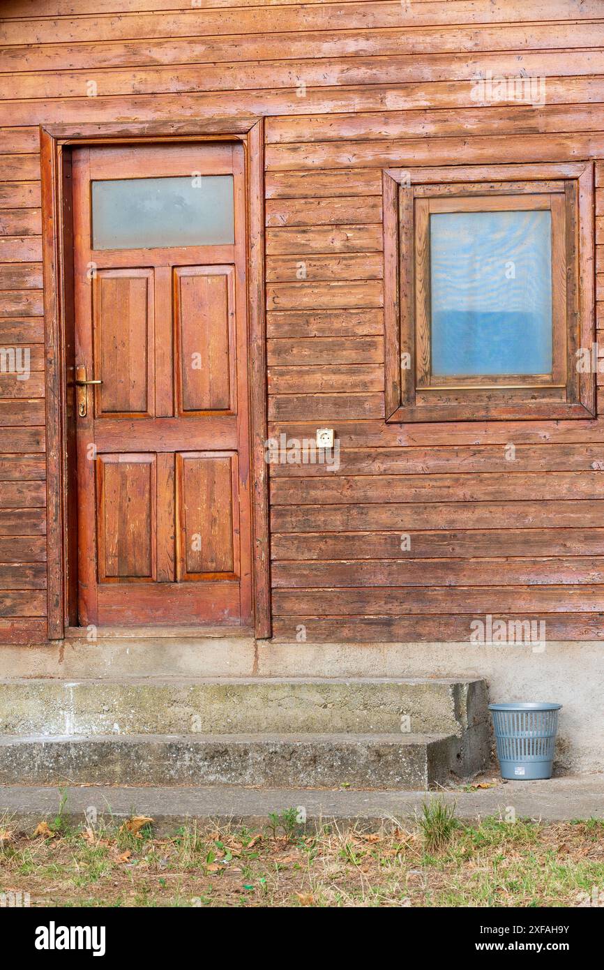 Forest Cabin House. Porta e telaio della finestra marrone di ingresso con tavole di legno e cestino in plastica per la spazzatura tramite scale di cemento. Foto Stock