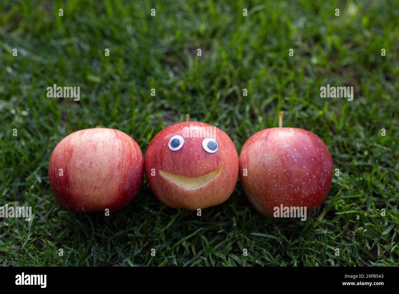 3 mele rosse su erba verde. una mela con un sorriso e gli occhi scolpiti su di essa. concetto positivo, scelta per essere felici. Stile di vita sano, vitamine naturali. V Foto Stock