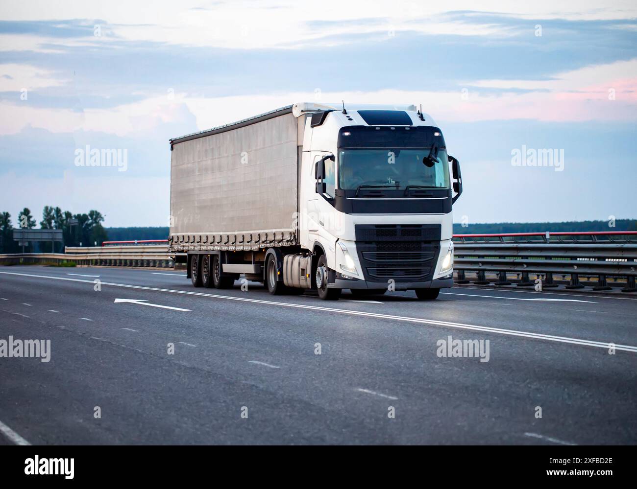 Un veicolo moderno con semirimorchio inclinabile trasporta il carico in autostrada di sera con i fari accesi contro il cielo. Concetto di trasposizione del carico Foto Stock