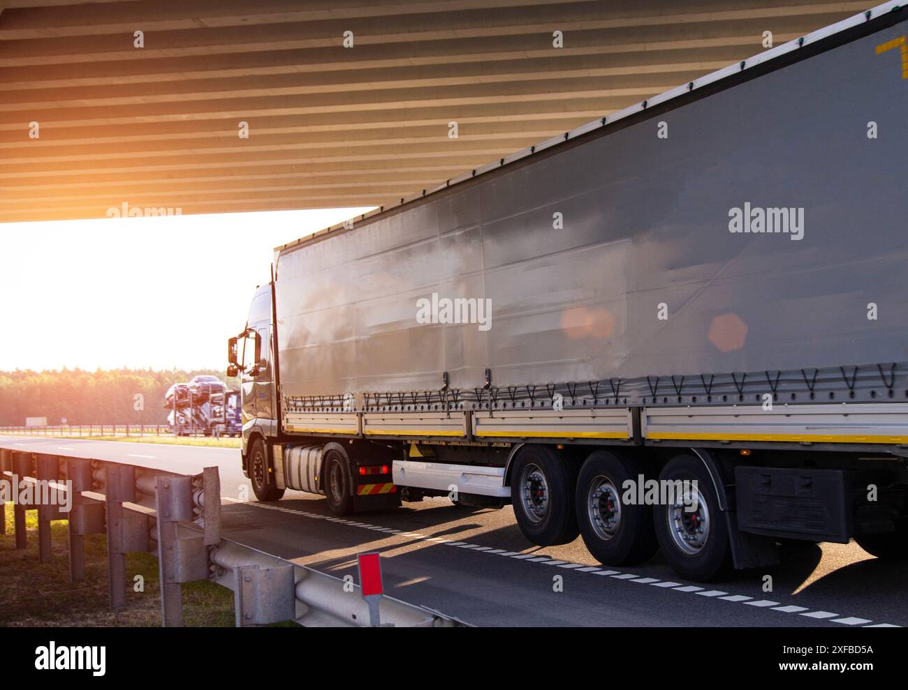 Un veicolo con semirimorchio inclinabile trasporta il carico sotto un ponte sullo sfondo del tramonto, copiando lo spazio per il testo Foto Stock