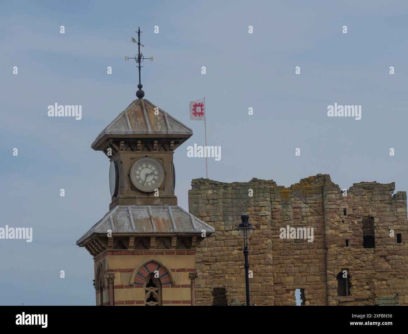 Una torre dell'orologio di fronte a una vecchia rovina di pietra con una bandiera ondulata sotto un cielo nuvoloso, tynemouth, inghilterra, Gran Bretagna Foto Stock