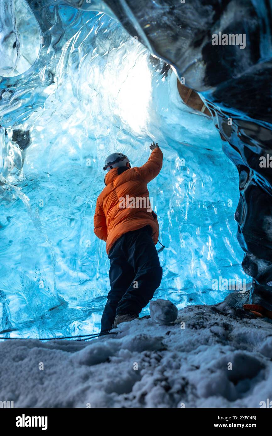 Un uomo con una giacca arancione si trova in una grotta blu. La grotta è piena di ghiaccio e l'uomo sta guardando verso il soffitto Foto Stock