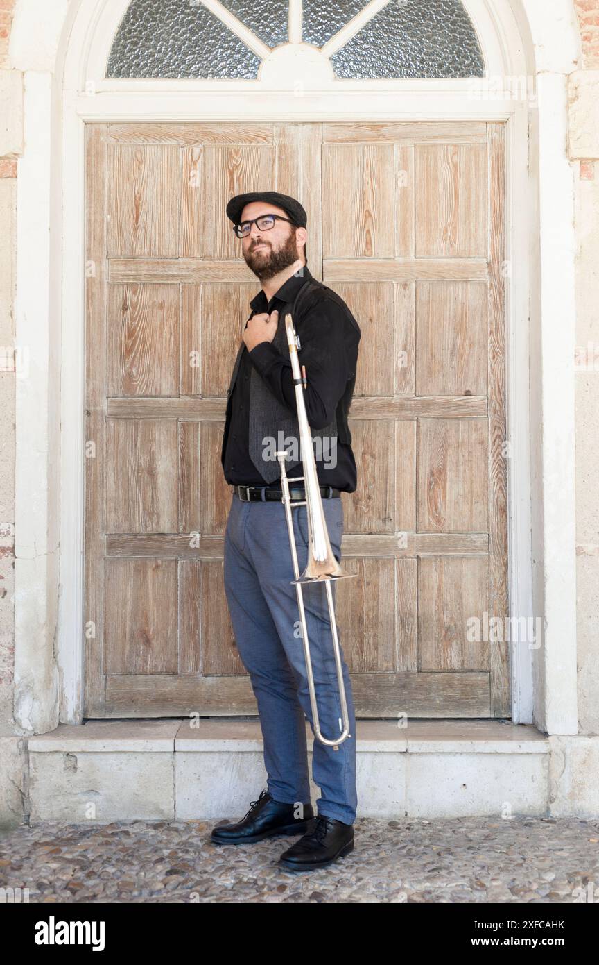 Musicista jazz con occhiali e barba che suona il trombone in piedi per strada. Vestito con un cappello inglese, una giacca, pantaloni blu. Dietro è un d di legno Foto Stock