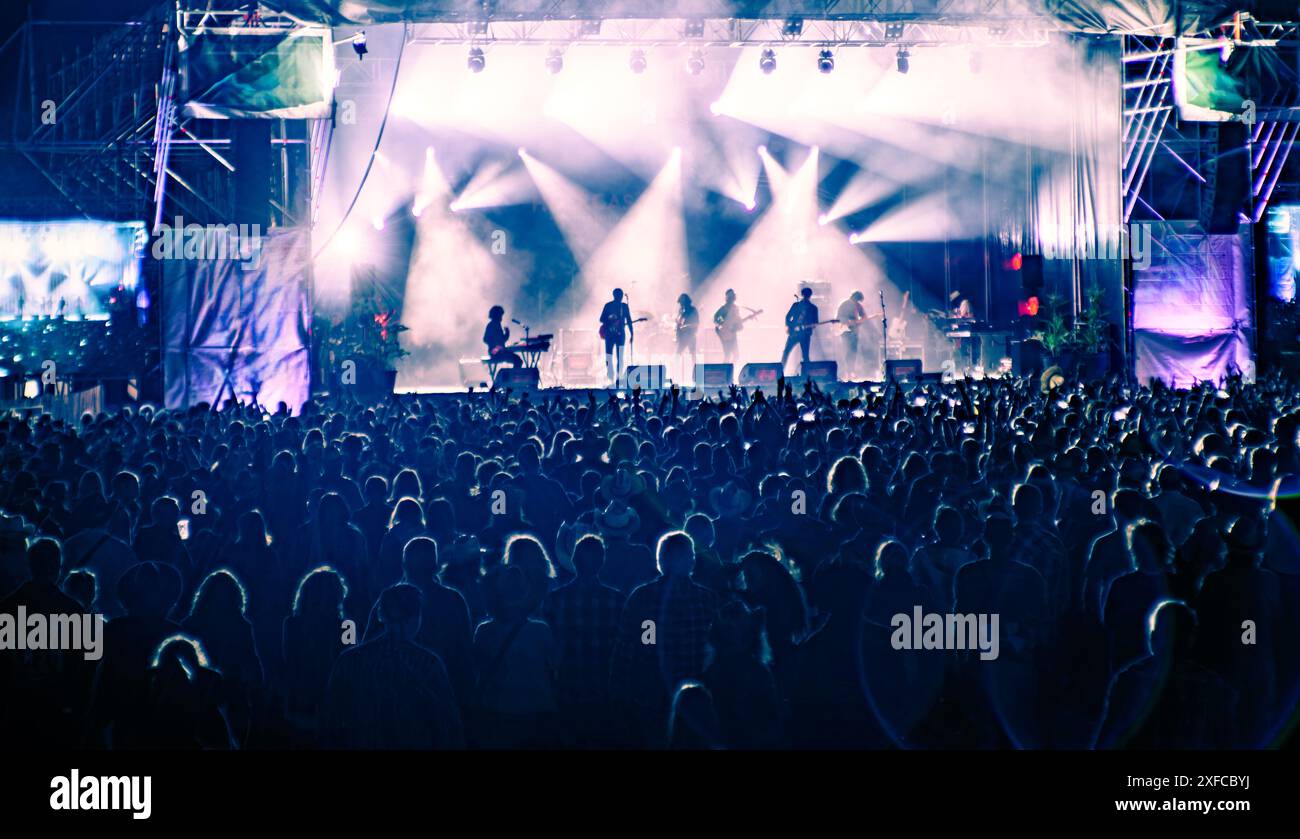 Una grande folla che si gode un concerto di musica dal vivo di sera. Il palco è illuminato da luci colorate, creando un'atmosfera elettrizzante. Foto Stock