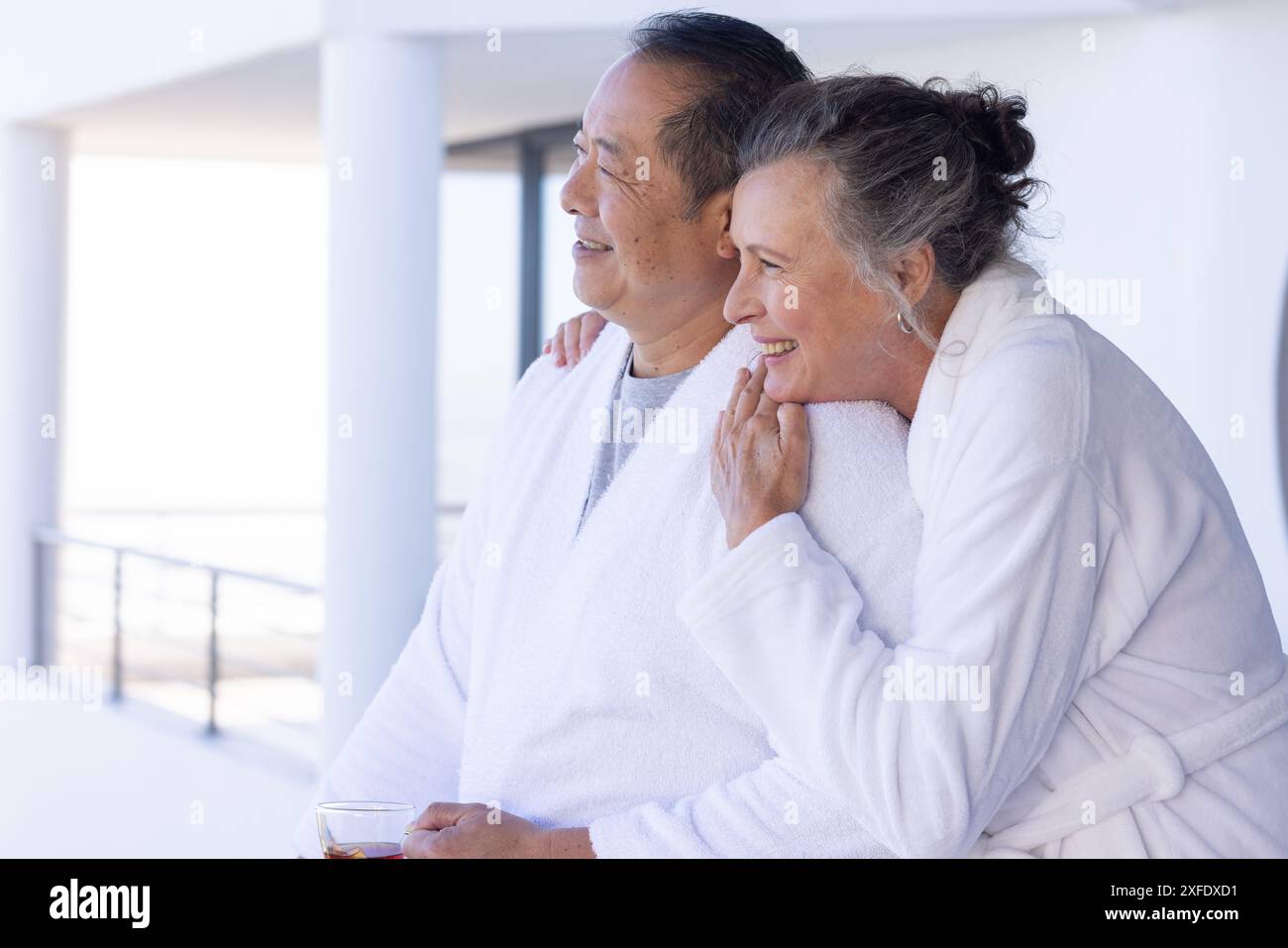 Una coppia anziana in accappatoi che si diverte in vacanza, abbracciando e sorridendo sul balcone Foto Stock