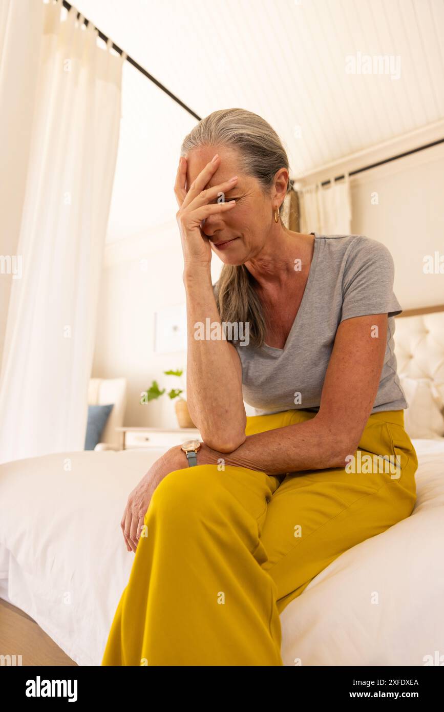 Seduta sul letto, donna matura con pantaloni gialli che sembra stressata e stanca Foto Stock