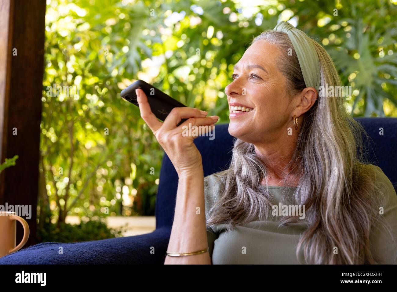 Donna matura sorridente che utilizza l'assistente vocale dello smartphone mentre si rilassa sulla sedia a casa Foto Stock