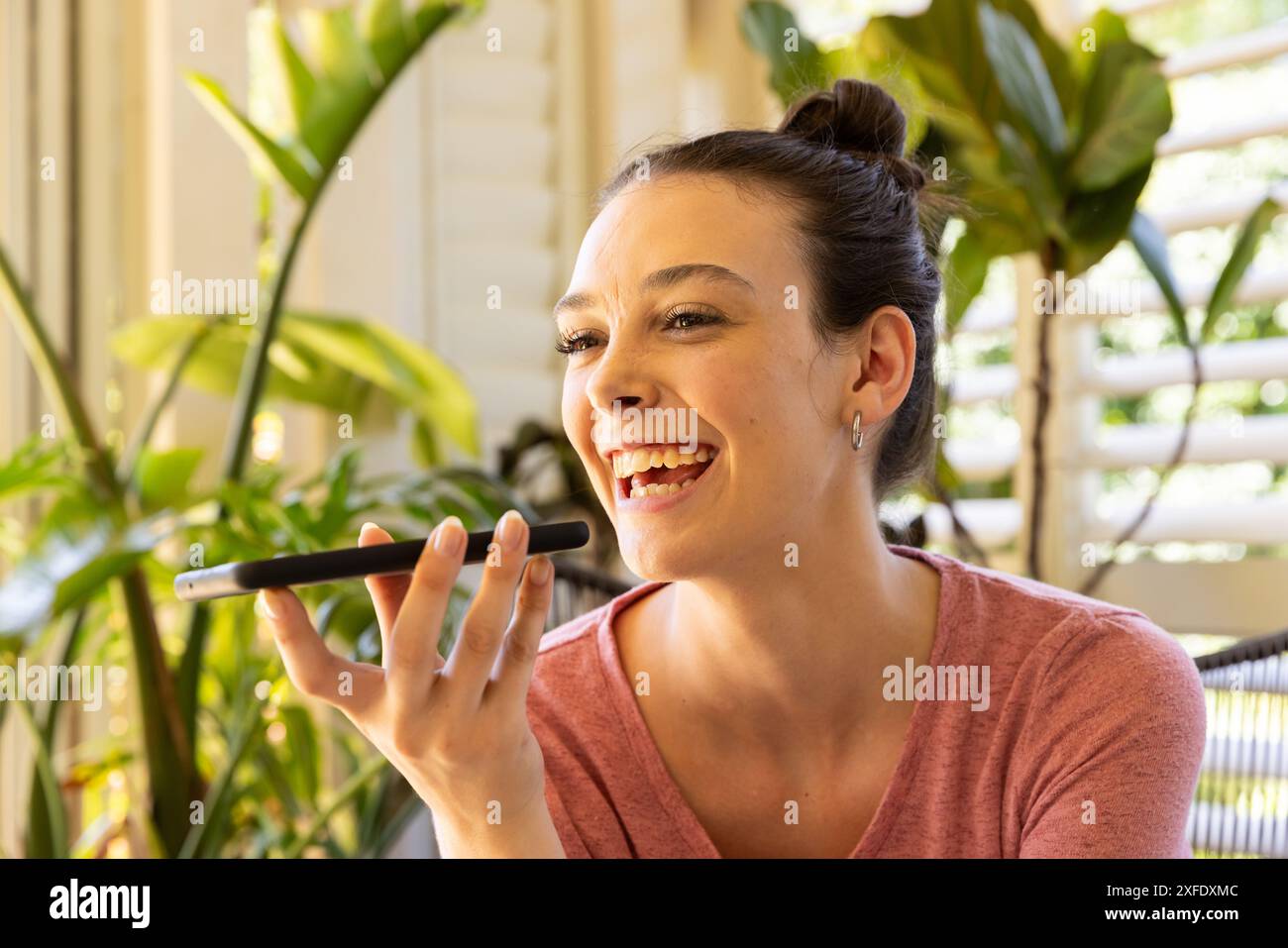 Donna sorridente che utilizza l'assistente vocale dello smartphone mentre si rilassa a casa con le piante Foto Stock