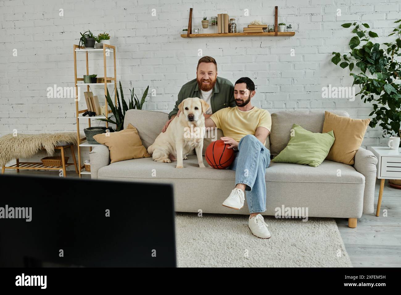 Due uomini con la barba si rilassano su un divano con il loro Labrador in un accogliente soggiorno. Foto Stock