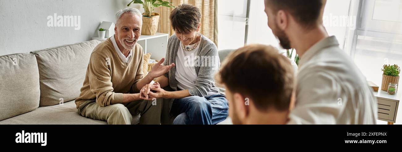 Una coppia gay siede con i genitori su un divano a casa loro, a fare una conversazione. Foto Stock