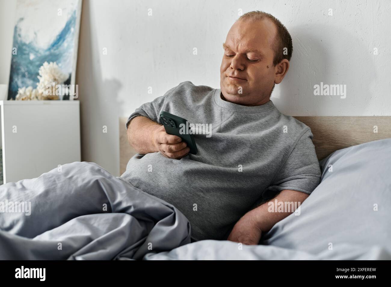Un uomo con l'inclusività si siede a letto, usando il suo smartphone, iniziando la sua giornata. Foto Stock