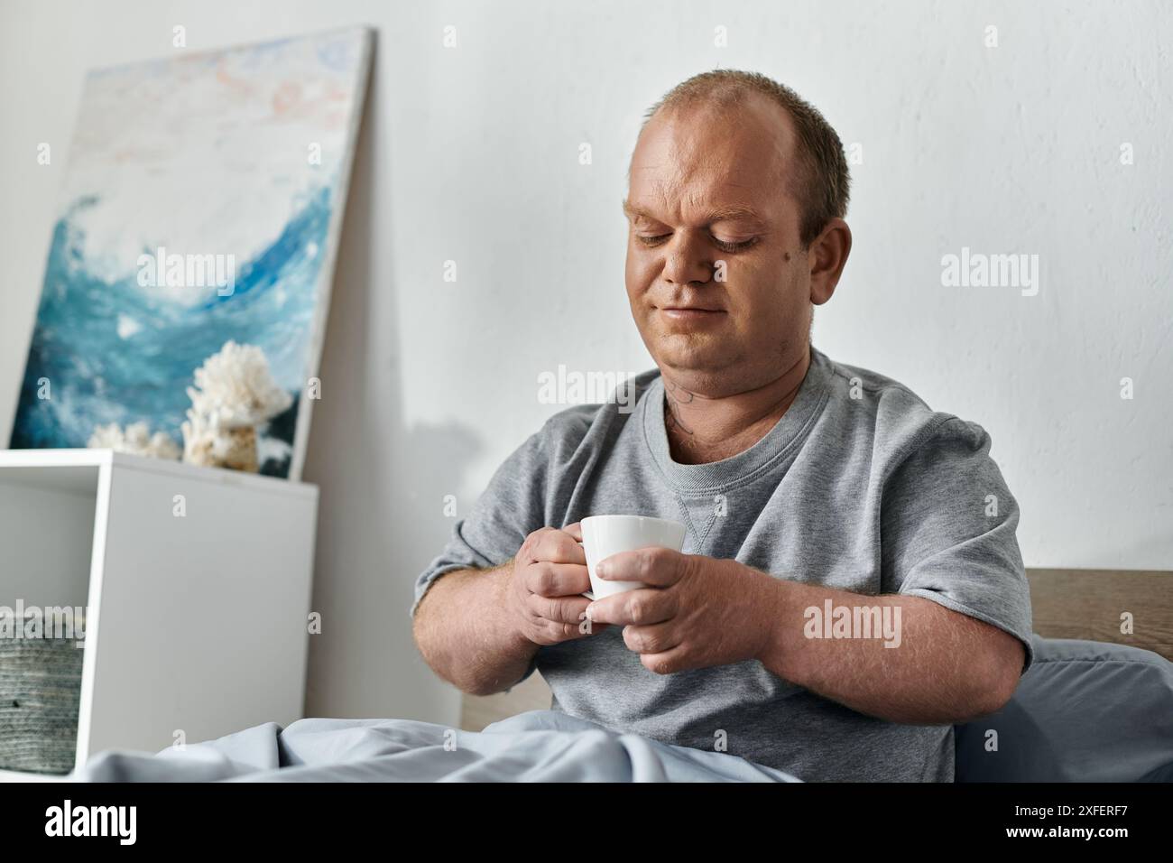 Un uomo con l'inclusività si siede a letto, tenendo in mano una tazza di caffè. Foto Stock