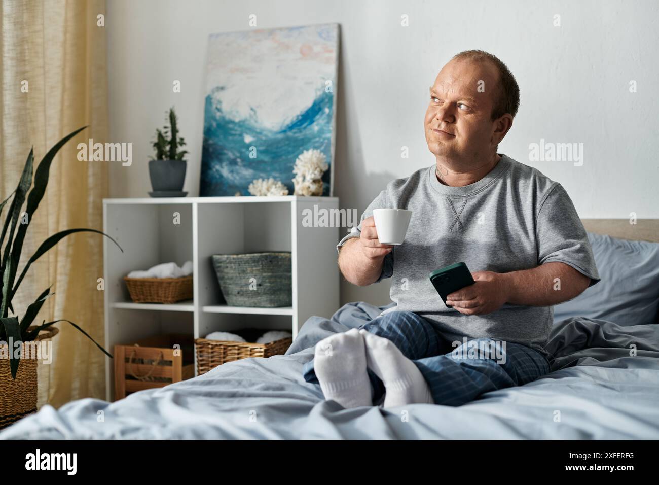 Un uomo con inclusività siede a letto, si gusta una tazza di caffè e guarda fuori dalla finestra, magari contemplando la sua giornata. Foto Stock