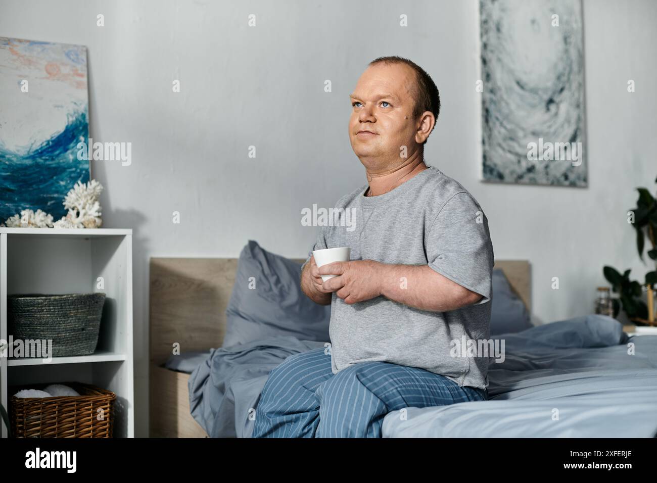 Un uomo con inclusività si siede sul suo letto la mattina, tenendo una tazza e guardando con attenzione fuori dall'inquadratura. Foto Stock