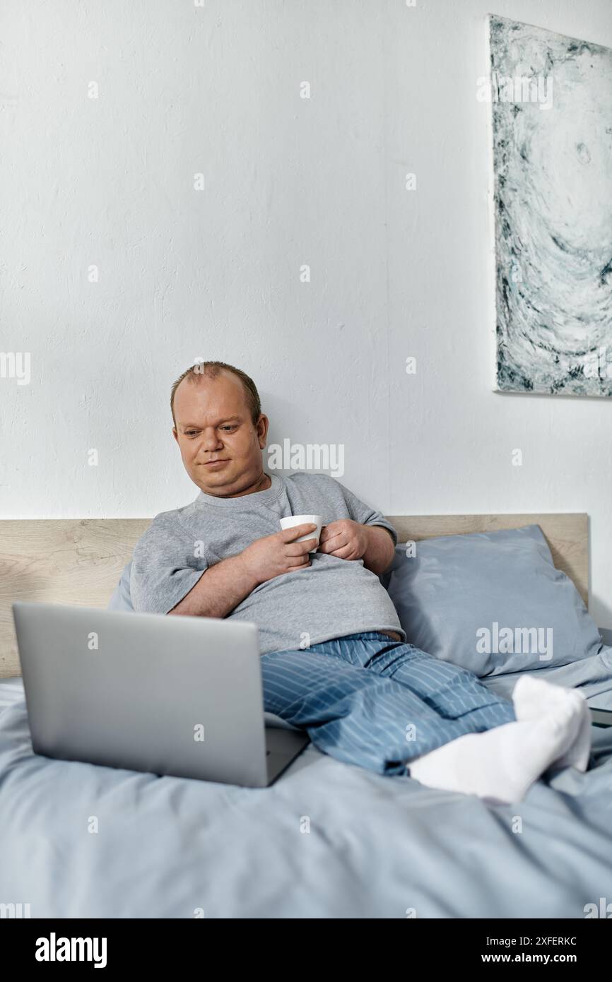 Un uomo con inclusività in pigiama si rilassa a letto, tenendo una tazza di caffè e guardando un computer portatile. Foto Stock
