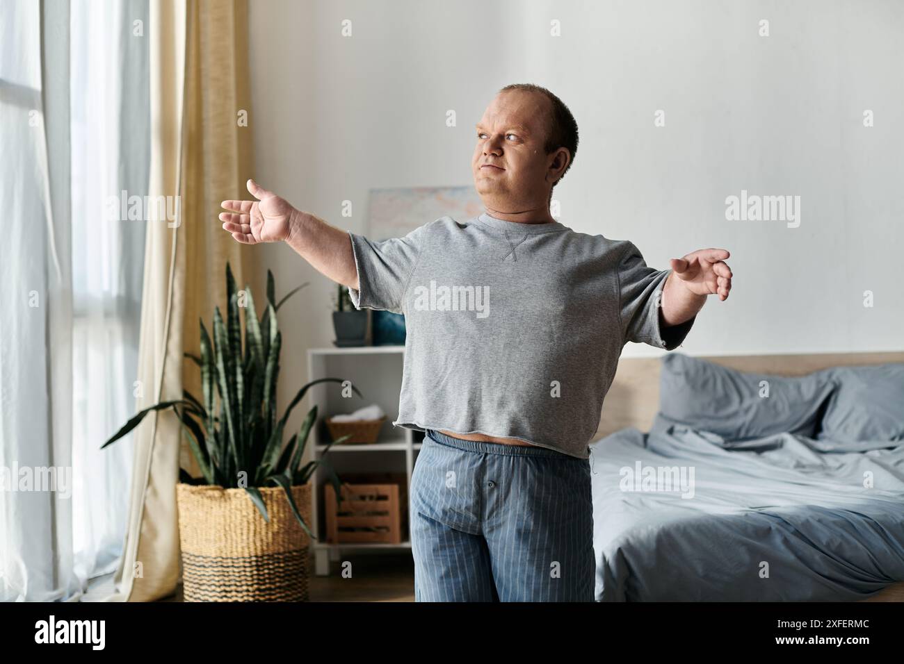 Un uomo con inclusività allunga le braccia nel pigiama mentre si trova vicino a una finestra. Foto Stock