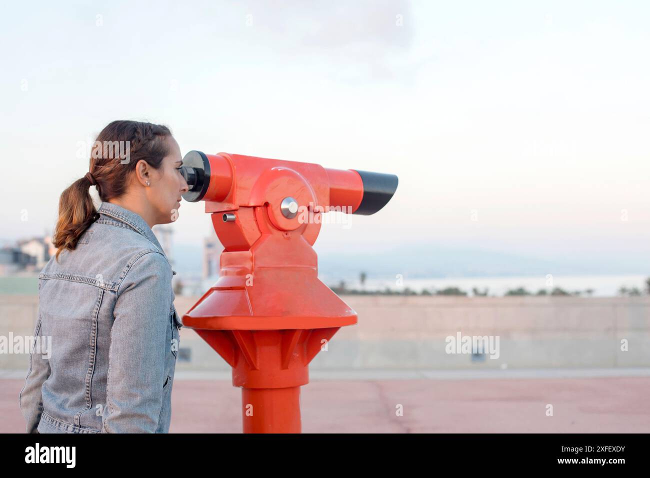 Una donna con giacca in denim guarda attraverso un telescopio rosso su un ponte di osservazione all'aperto, con strutture metalliche e un campo erboso sullo sfondo. Foto Stock