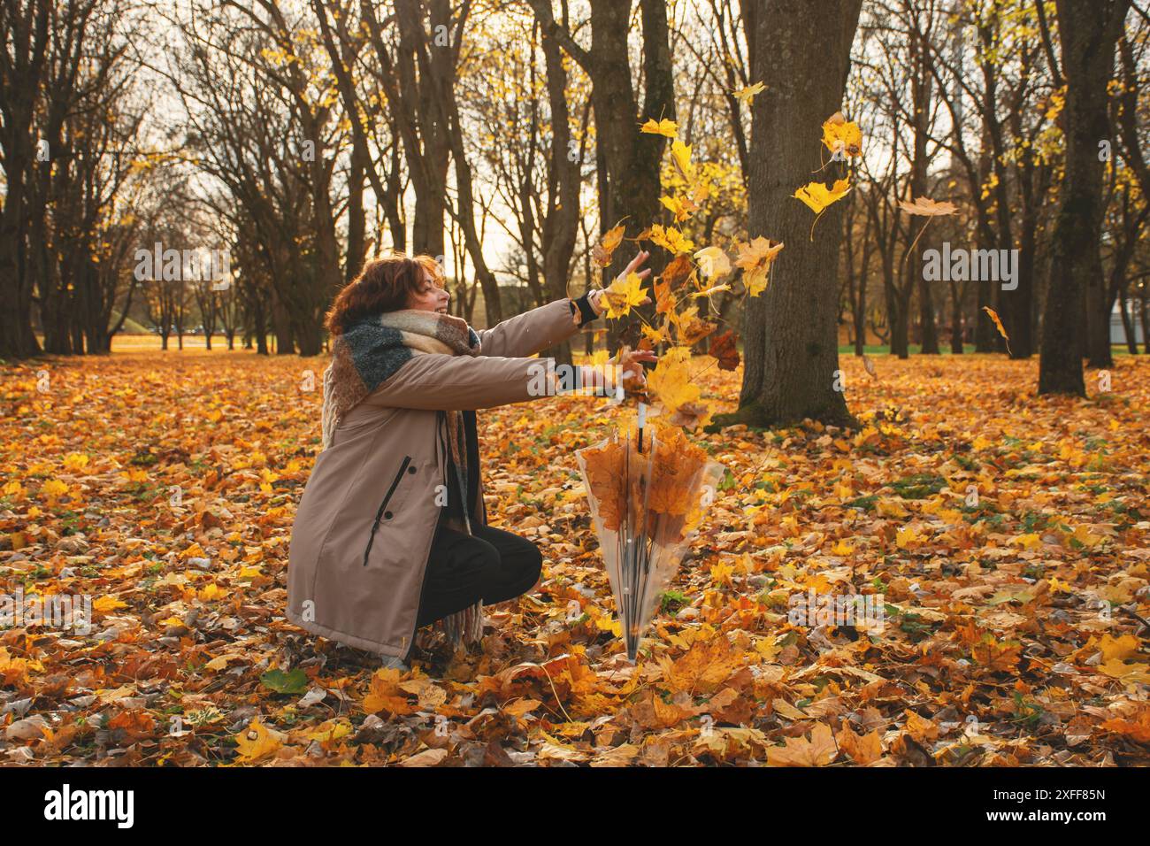 una donna di 50 anni cammina nel parco in autunno in una calda giornata di sole, cattura le foglie cadenti con le mani e ridendo Foto Stock