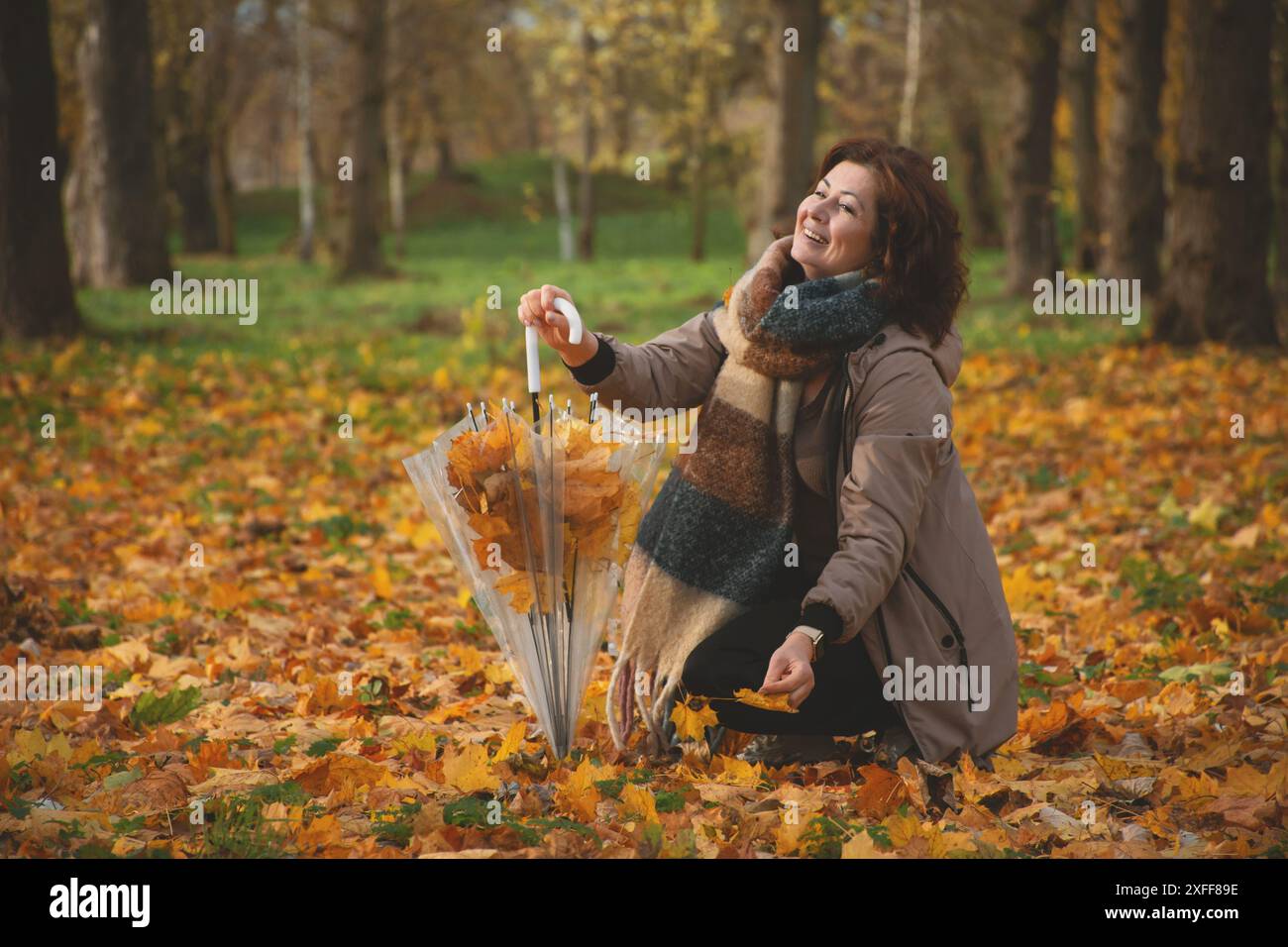 una donna di 50 anni cammina nel parco in autunno, raccoglie le foglie cadenti in un ombrello e ride Foto Stock