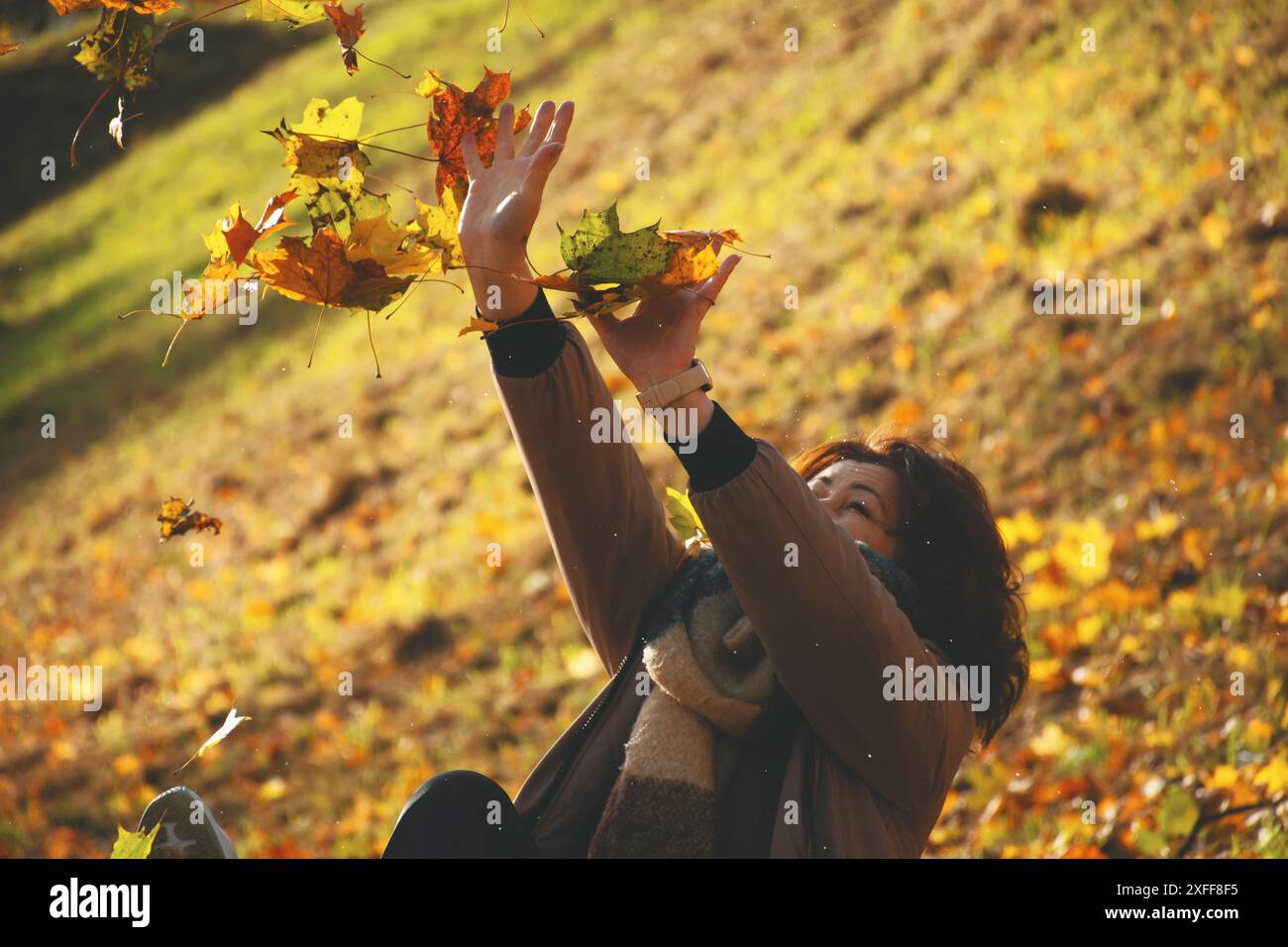 Una donna di 50 anni si protegge dalle foglie cadenti con le mani, gioca con le foglie in autunno nel parco, in una calda giornata di sole, Foto Stock