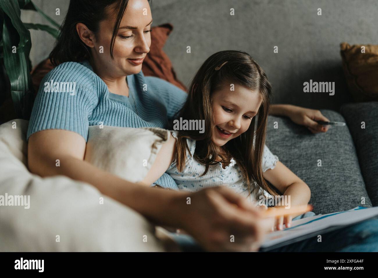 Madre che assiste la figlia felice a fare i compiti mentre si siede sul divano a casa Foto Stock