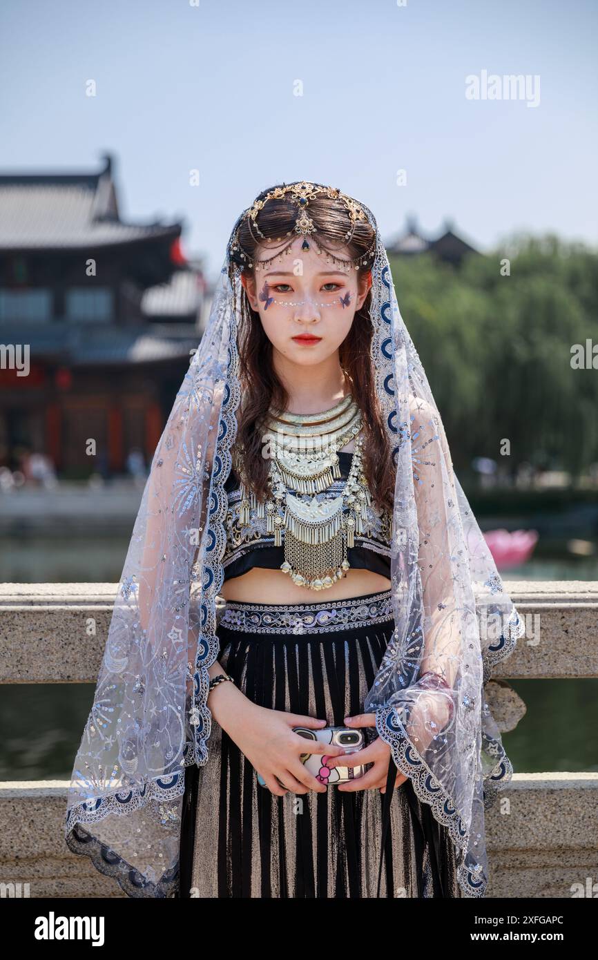 Un'affascinante donna asiatica adorna un intricato abbigliamento tradizionale, accostato a uno sfondo sereno caratterizzato da un ponte Foto Stock