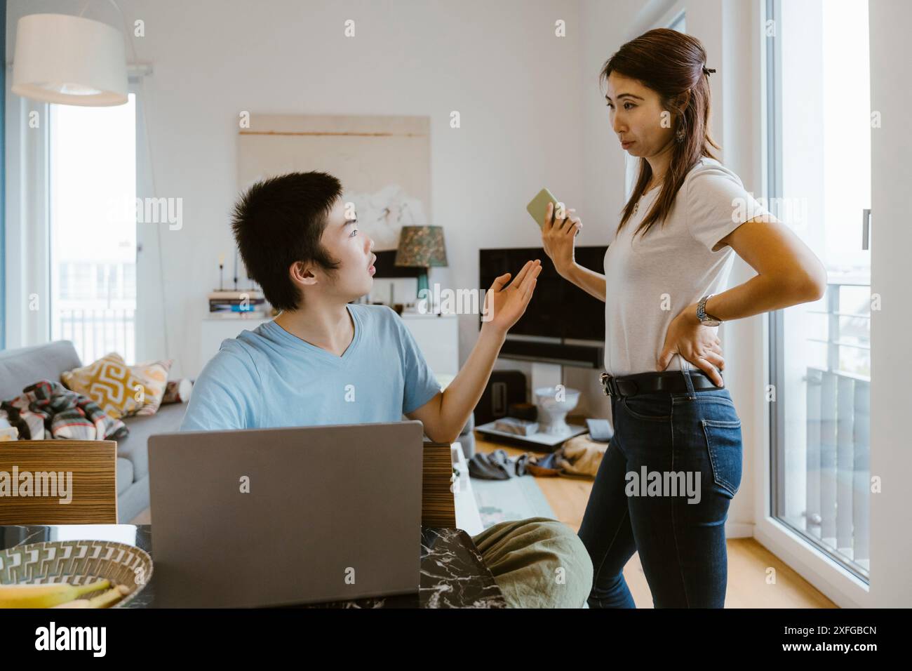 Madre con la mano sull'anca che prende lo smartphone dal figlio mentre studia a casa Foto Stock