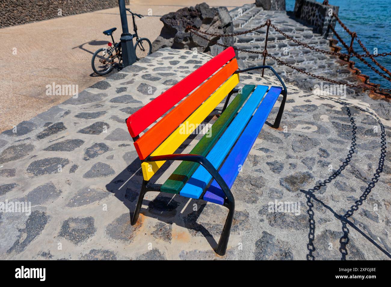 Panca colorata arcobaleno su passerella in pietra. La panchina colorata dell'arcobaleno si trova su un passaggio in pietra accanto a un corpo d'acqua. Il banco è circondato da una maglia a catena Foto Stock