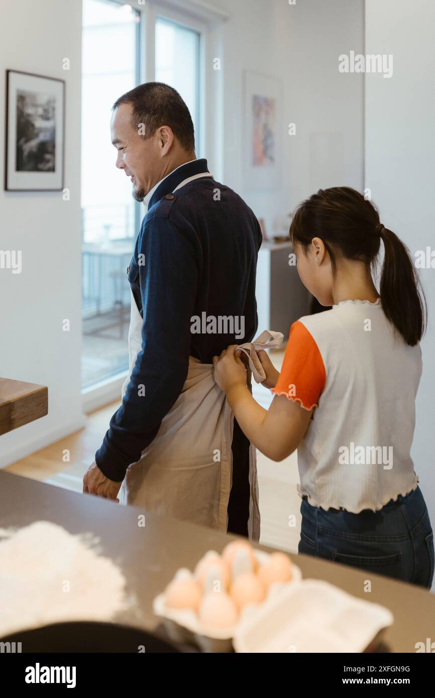 Ragazza che aiuta il padre mentre indossa il grembiule in cucina a casa Foto Stock