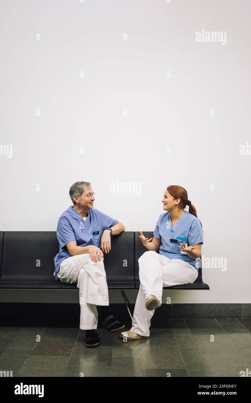 Tirocinante femminile sorridente che discute con il medico di sesso maschile anziano seduto sulla sedia contro il muro bianco in ospedale Foto Stock