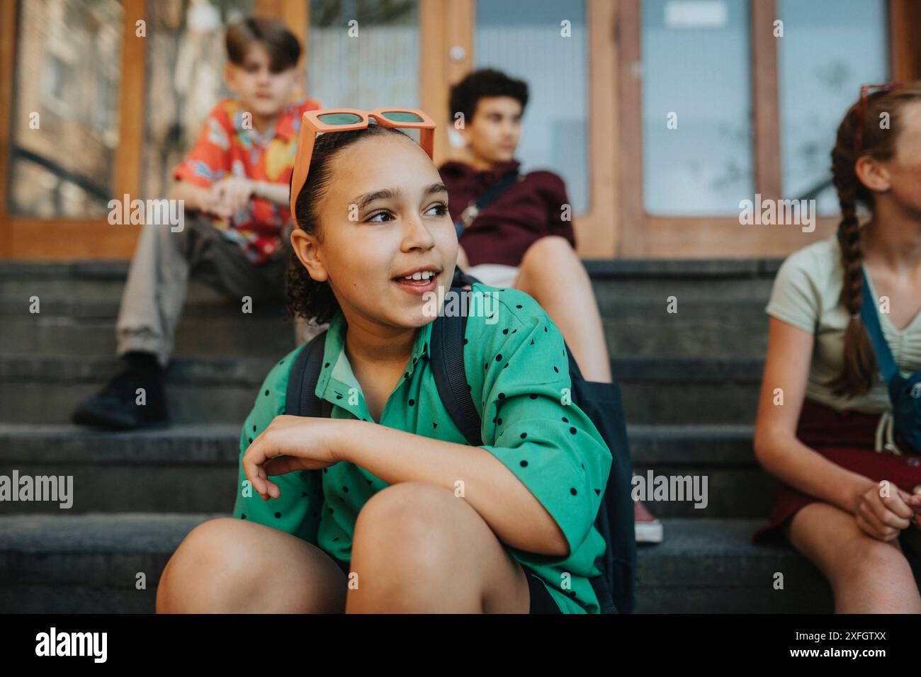 Ragazza che indossa gli occhiali da sole seduta con gli amici sulle scale dell'ingresso dell'edificio scolastico Foto Stock