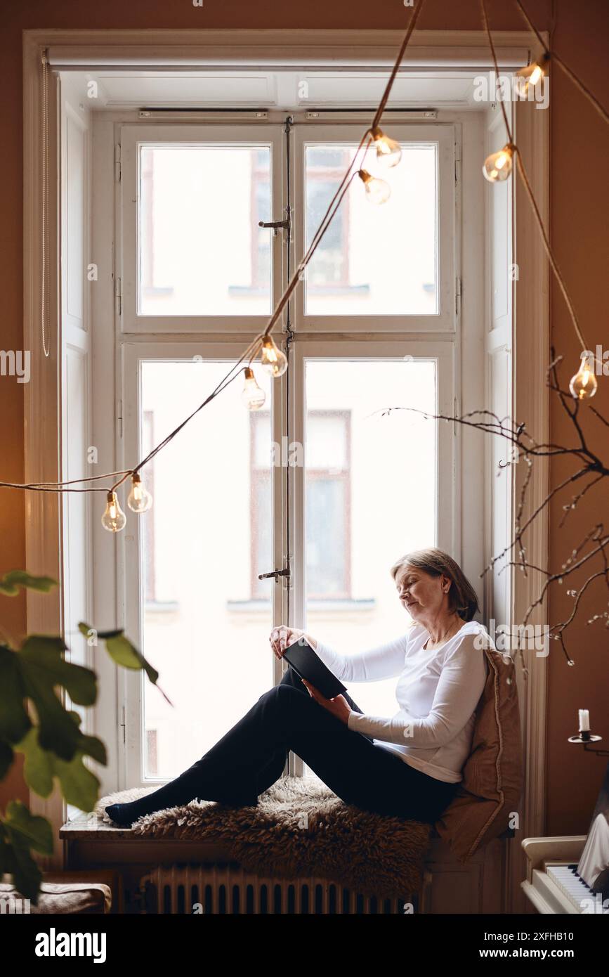 Donna anziana che legge un libro seduto sul davanzale della finestra a casa Foto Stock