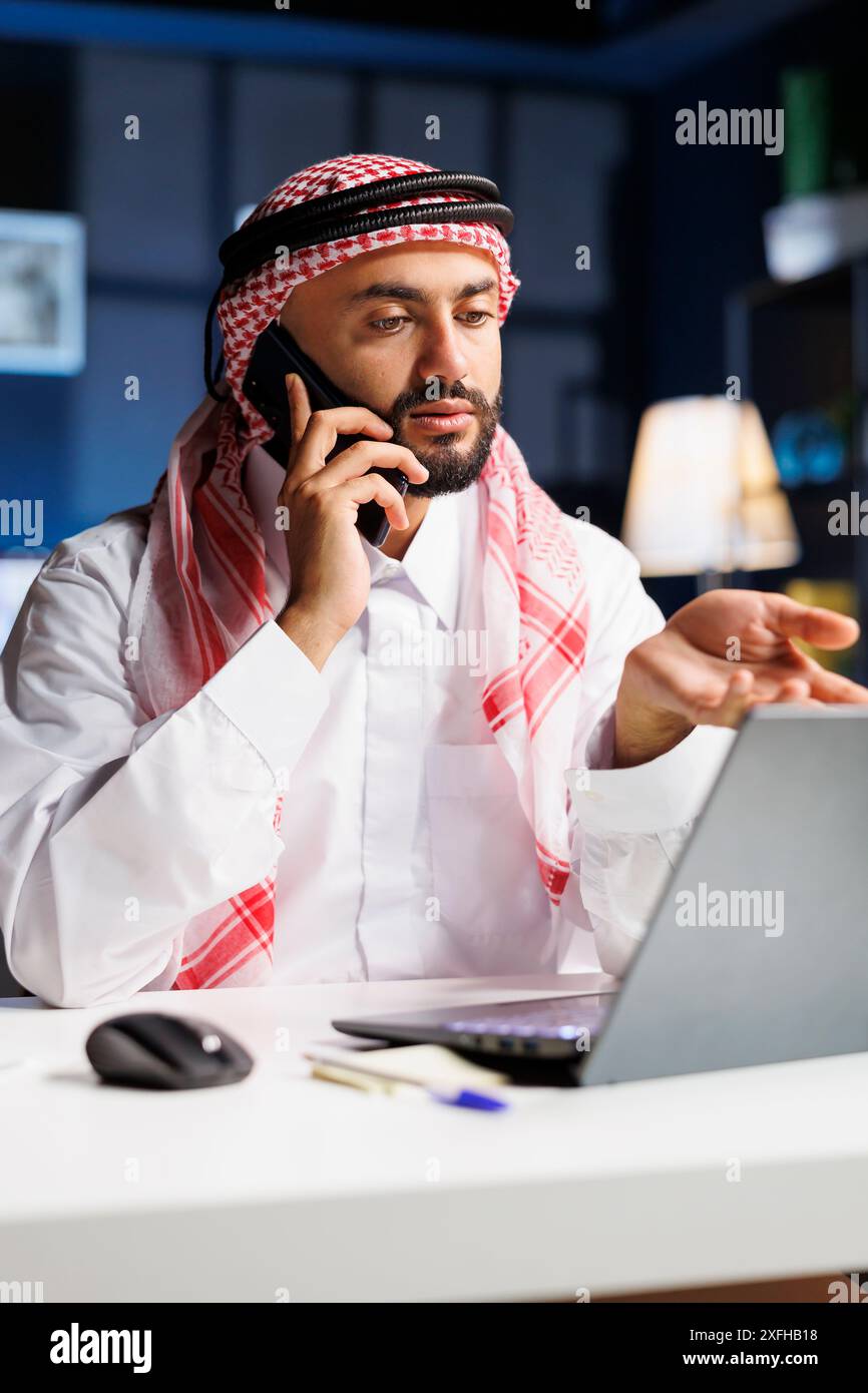 Foto dettagliata di un uomo musulmano che si concentra su una conversazione telefonica mentre controlla il suo portatile. Il ragazzo arabo che abbraccia la tecnologia dimostra professionalità e forti capacità di comunicazione. Foto Stock
