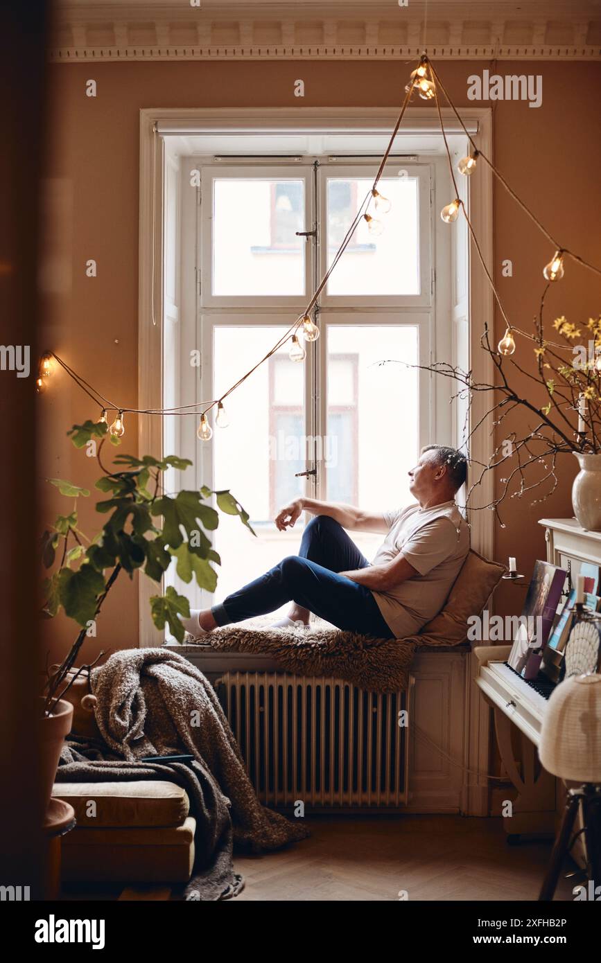 Vista laterale di un uomo maturo seduto sul davanzale della finestra a casa Foto Stock