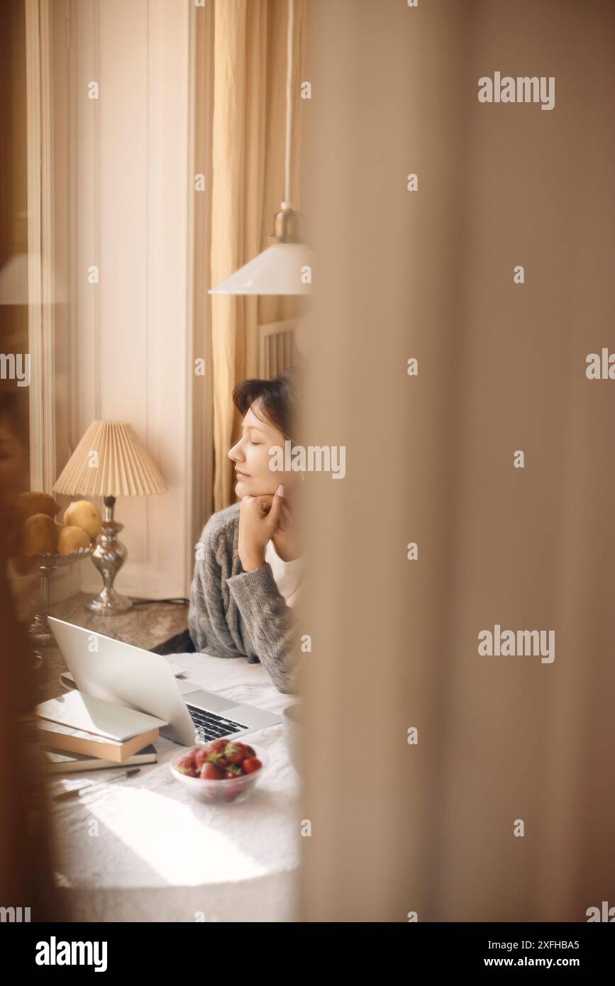 Giovane donna con la mano sul mento seduta a tavola in casa Foto Stock