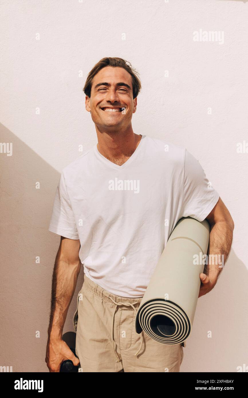 Ritratto di un uomo sorridente che tiene il tappetino da yoga arrotolato e si trova vicino al muro Foto Stock