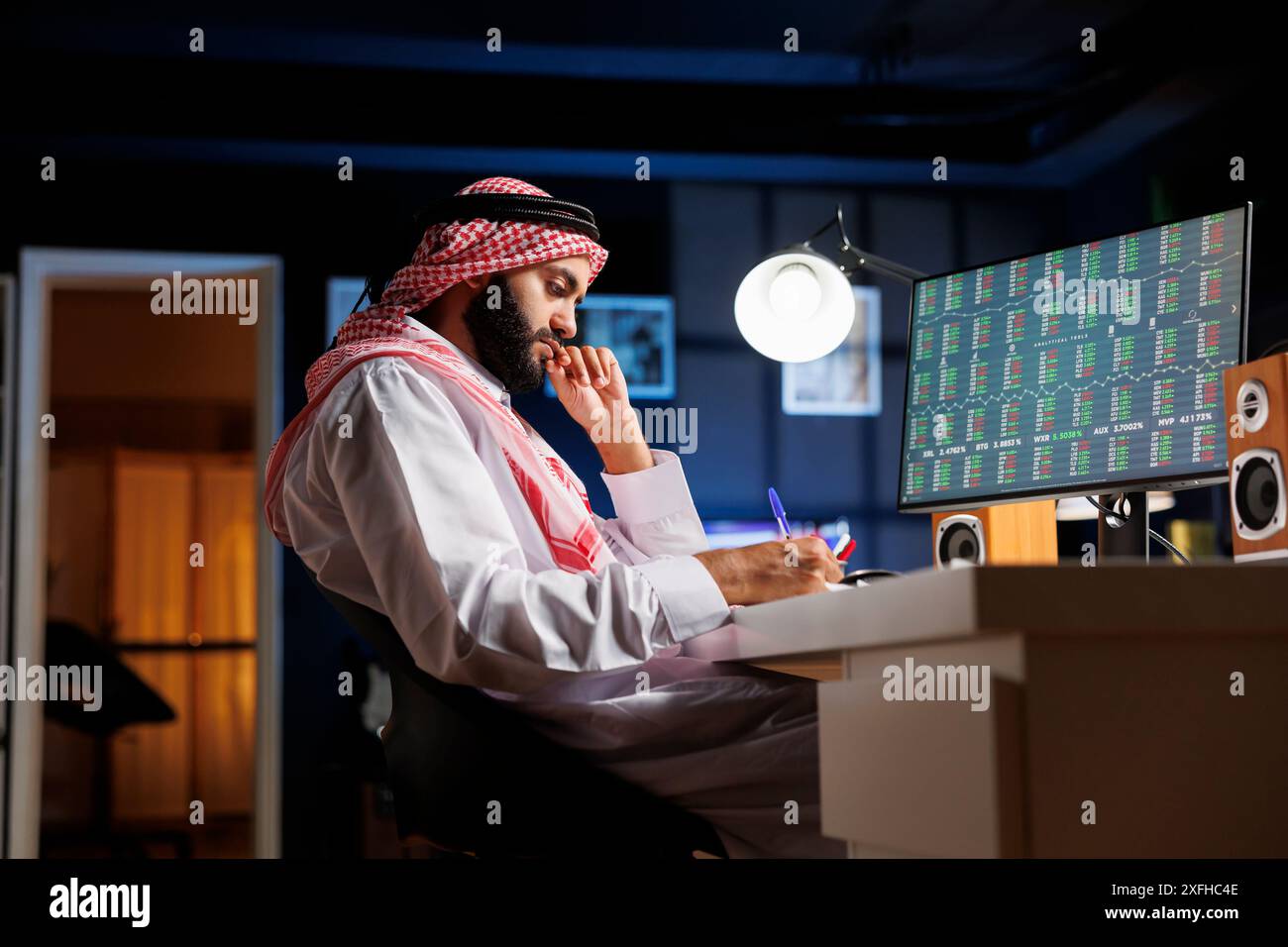 Il tizio del Medio Oriente sedeva alla sua postazione di lavoro, studiando e prendendo appunti dal suo computer desktop. Questa immagine mostra un uomo arabo con un blocco note che sta eseguendo il brainstorming e utilizza un computer. Foto Stock