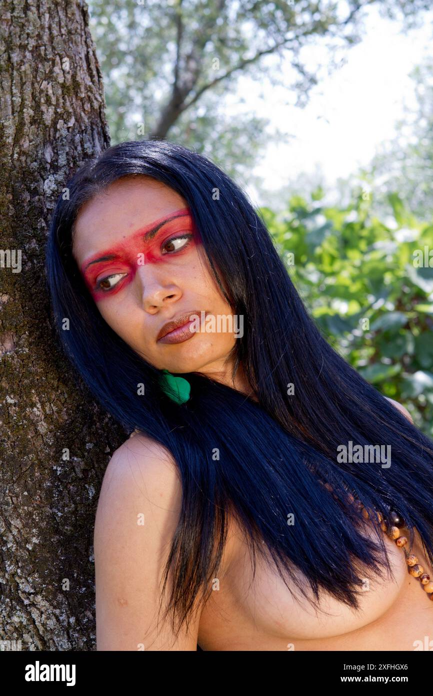 Ritratto di una donna indigena brasiliana con tradizionale pittura facciale, accovacciata in un lussureggiante ambiente forestale. Foto Stock