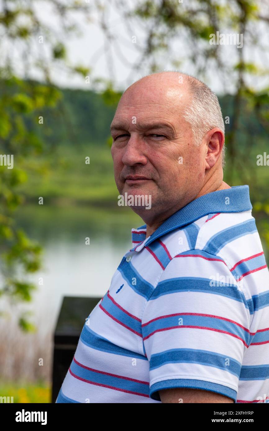 Un uomo calvo di mezza età con una camicia a righe si trova vicino a un lago. Foto Stock