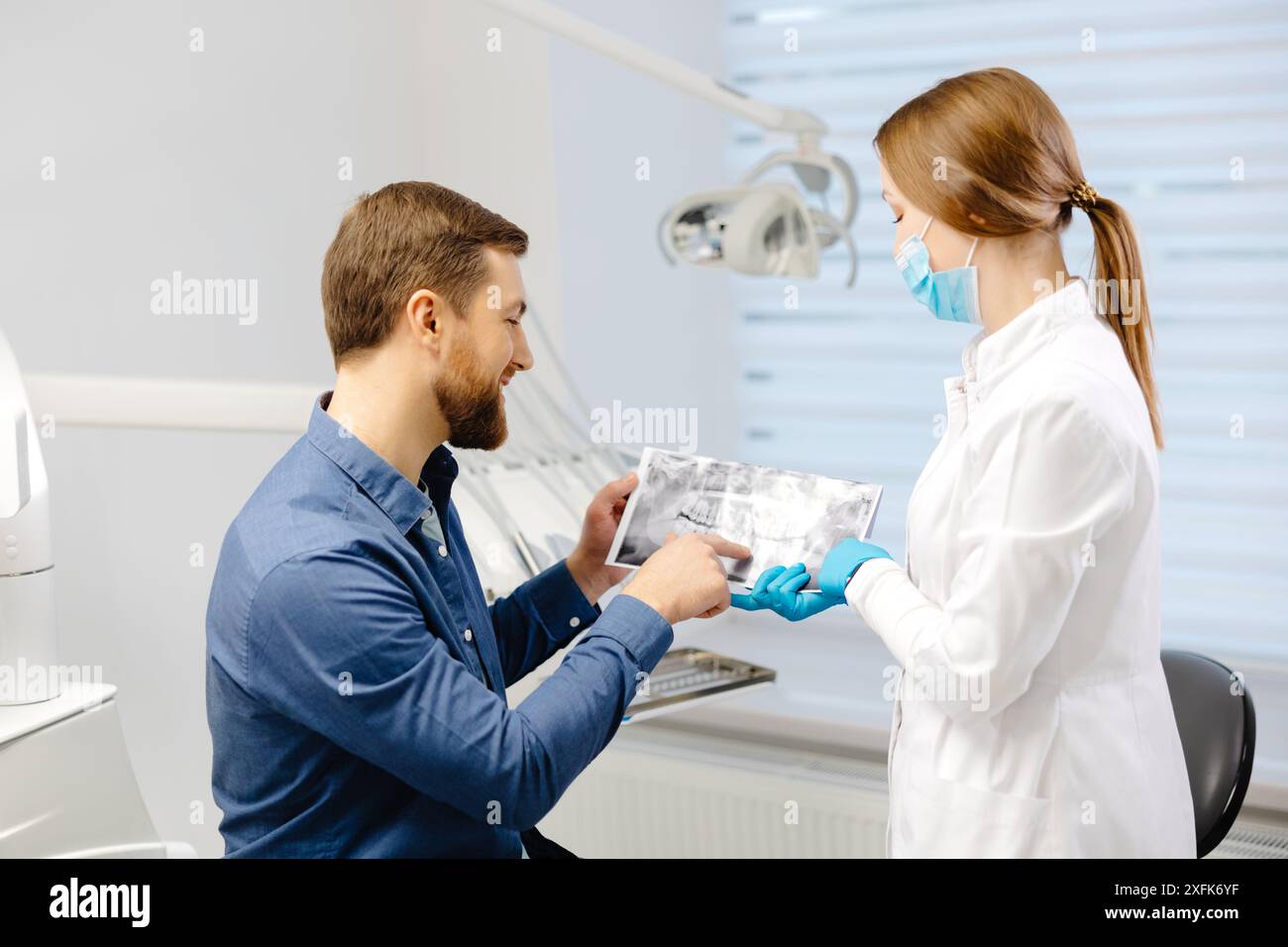 Giovane uomo attraente in visita al dentista, seduto su una sedia dentale presso una moderna clinica leggera. Una giovane dentista che tiene in mano una imagewoman a raggi X. Foto Stock