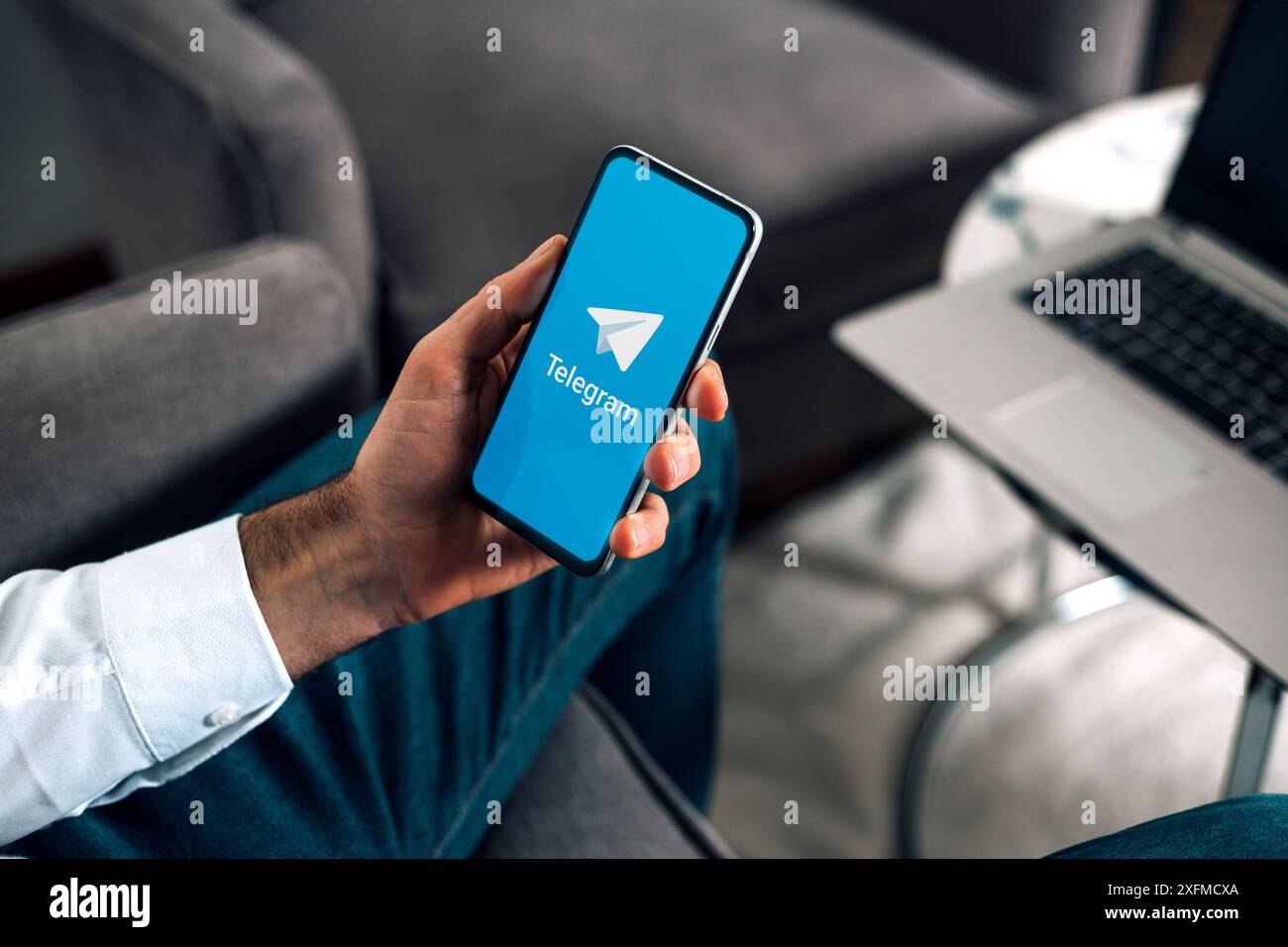 Uomo con un telefono cellulare che mostra l'app telegramma sullo schermo. Uomo sul posto di lavoro con laptop e smartphone in mano. ROSARIO, ARGENTINA - 3 LUGLIO 2024. Foto Stock