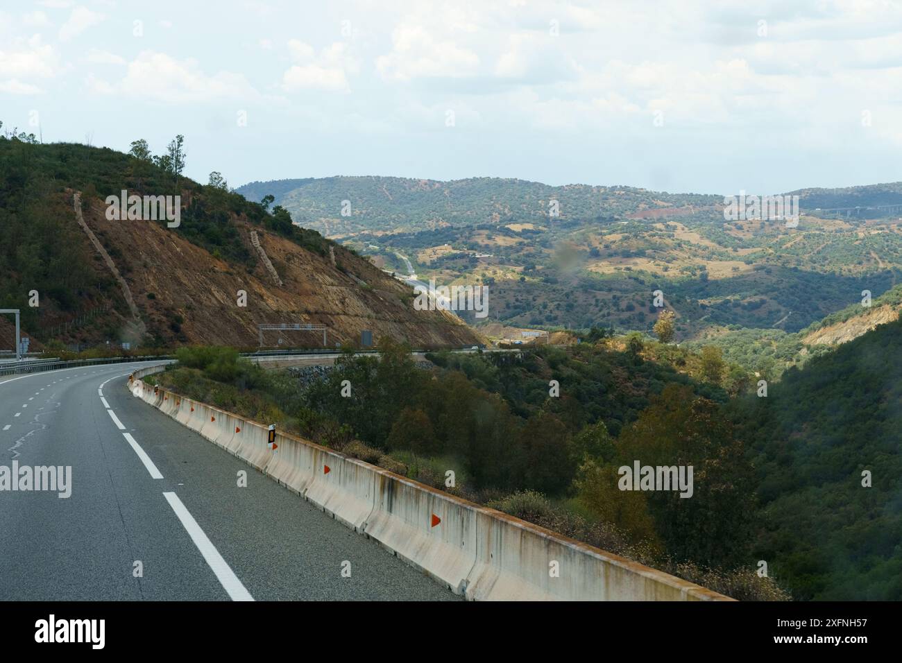 Una strada si snoda attraverso dolci colline, delimitata da una barriera di cemento. Il cielo limpido con nuvole mostruose illumina il paesaggio verdeggiante. Foto Stock