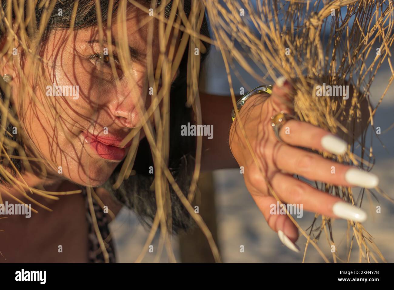 Questa immagine presenta un ritratto creativo di una donna su una spiaggia, sbirciando attraverso un ombrellone di paglia. Foto Stock
