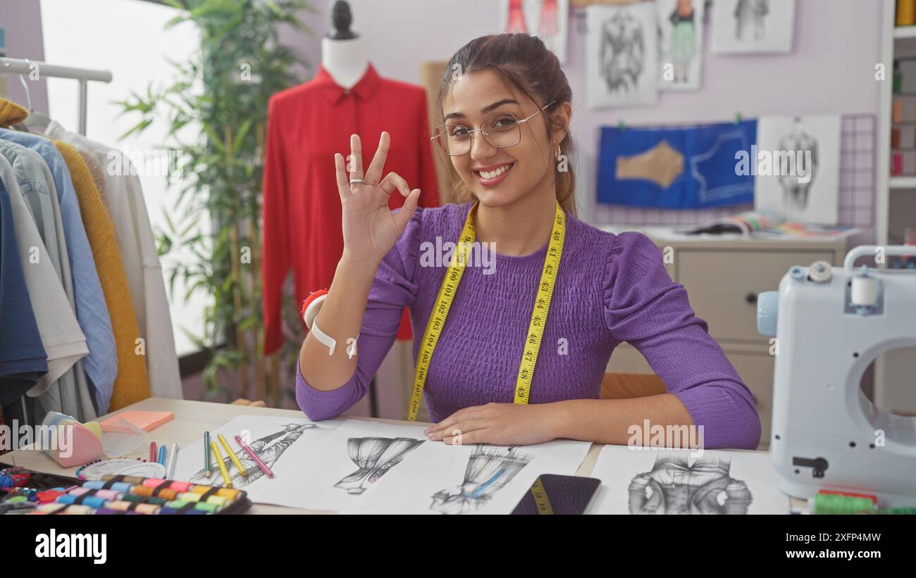 Una giovane donna ispanica sorridente che gestiva "ok" in una vivace sartoria con schizzi, tessuti e una macchina da cucire. Foto Stock