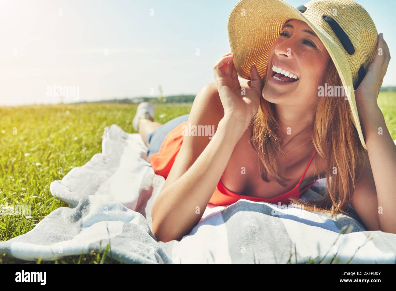 Donna, felice e rilassata sull'erba all'aperto per vacanze, sole e benessere con cielo blu o coperta. Donna, cappello e sorriso nel parco di New York Foto Stock