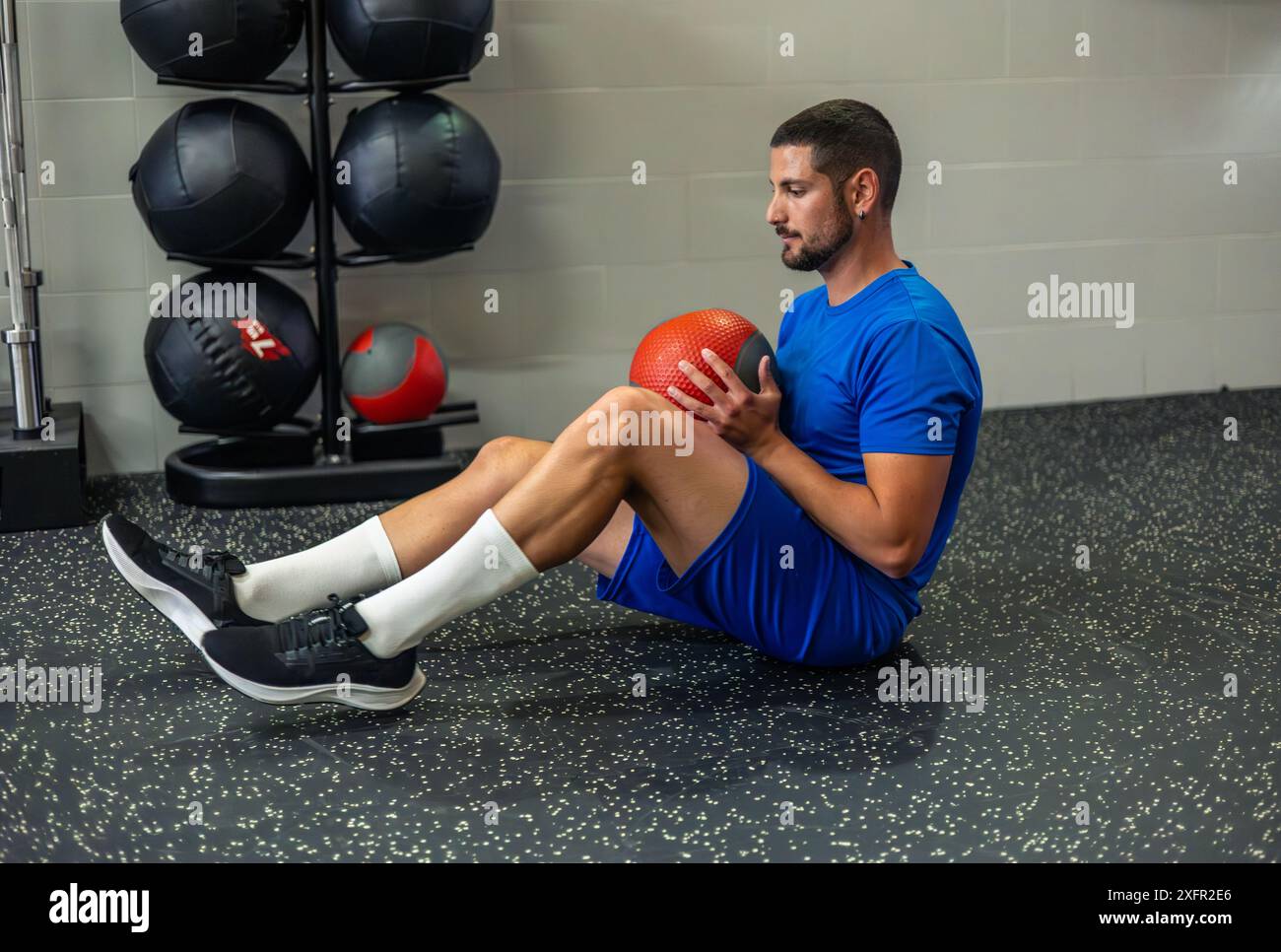 il giovane atleta è seduto sul pavimento della palestra eseguendo esercizi di base con una palla medica. Questo allenamento si concentra sulla forza e la stabilità del core, showcasi Foto Stock