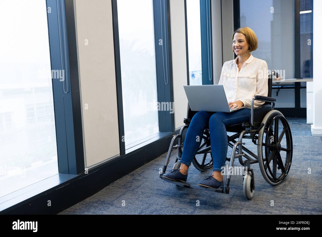 Donna sorridente in sedia a rotelle che utilizza un computer portatile, lavora in un ufficio moderno, copia spazio Foto Stock