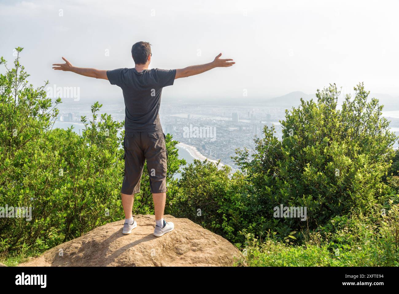 Uomo che alza le mani e gode di una splendida vista e di aria fresca sulla cima della montagna tra cespugli verdi. Una città costiera è visibile sullo sfondo. Foto Stock